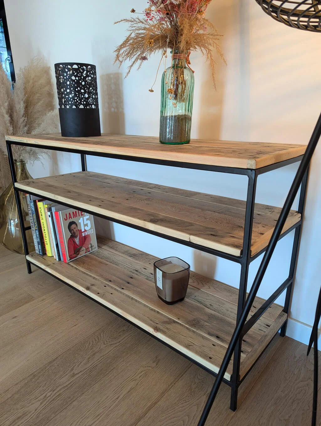 Étagère Louise en bois recyclé et métal noir, décorée avec un vase en verre rempli de fleurs séchées, une bougie et des livres de cuisine, apportant une touche industrielle et chaleureuse à l’intérieur.